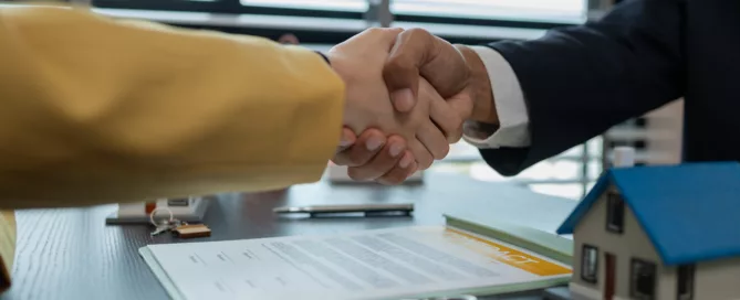 Two people shaking hands over a contract with a model house and keys on the table, symbolizing a real estate agreement.